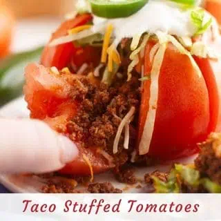 A close-up of a tomato stuffed with seasoned ground beef, shredded cheese, lettuce, and sour cream, with a finger pulling away a tomato slice.