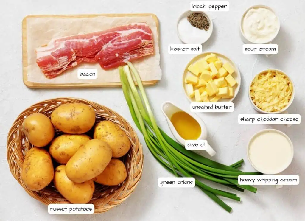 A variety of ingredients are arranged on a countertop, including russet potatoes, bacon, green onions, olive oil, unsalted butter, black pepper, kosher salt, sour cream, cheddar cheese, and heavy cream.