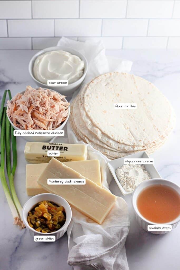 Ingredients for a recipe arranged on a countertop: flour tortillas, sour cream, cooked rotisserie chicken, butter, all-purpose cream, Monterey Jack cheese, chicken broth, green chiles, and green onions.
