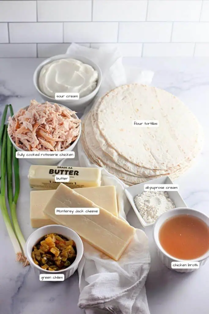 Ingredients for a recipe arranged on a countertop: flour tortillas, sour cream, cooked rotisserie chicken, butter, all-purpose cream, Monterey Jack cheese, chicken broth, green chiles, and green onions.