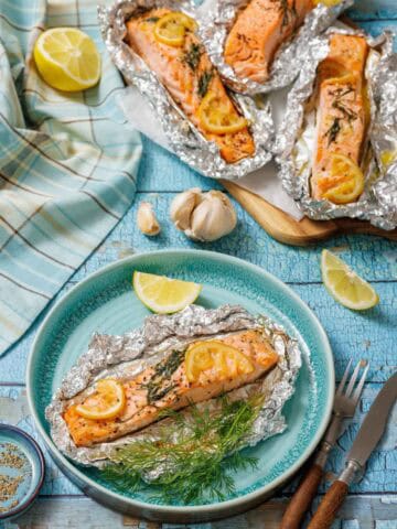 A serving of baked lemon-dill salmon in foil on a turquoise plate with lemon wedges and fresh dill. In the background, a baking sheet holds three more salmon foil packets, next to a blue and white checkered cloth and garlic cloves on a rustic blue wooden surface.