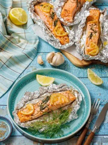 A serving of baked lemon-dill salmon in foil on a turquoise plate with lemon wedges and fresh dill. In the background, a baking sheet holds three more salmon foil packets, next to a blue and white checkered cloth and garlic cloves on a rustic blue wooden surface.