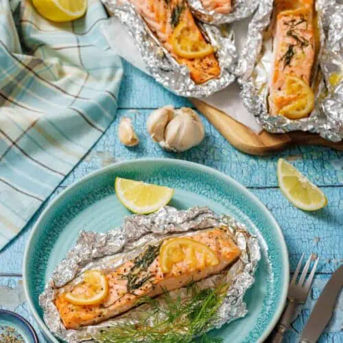 A serving of baked lemon-dill salmon in foil on a turquoise plate with lemon wedges and fresh dill. In the background, a baking sheet holds three more salmon foil packets, next to a blue and white checkered cloth and garlic cloves on a rustic blue wooden surface.