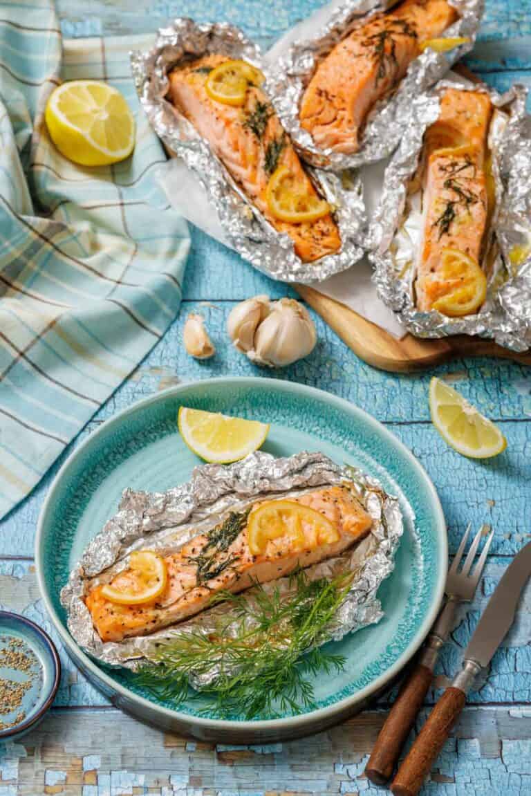 A serving of baked lemon-dill salmon in foil on a turquoise plate with lemon wedges and fresh dill. In the background, a baking sheet holds three more salmon foil packets, next to a blue and white checkered cloth and garlic cloves on a rustic blue wooden surface.