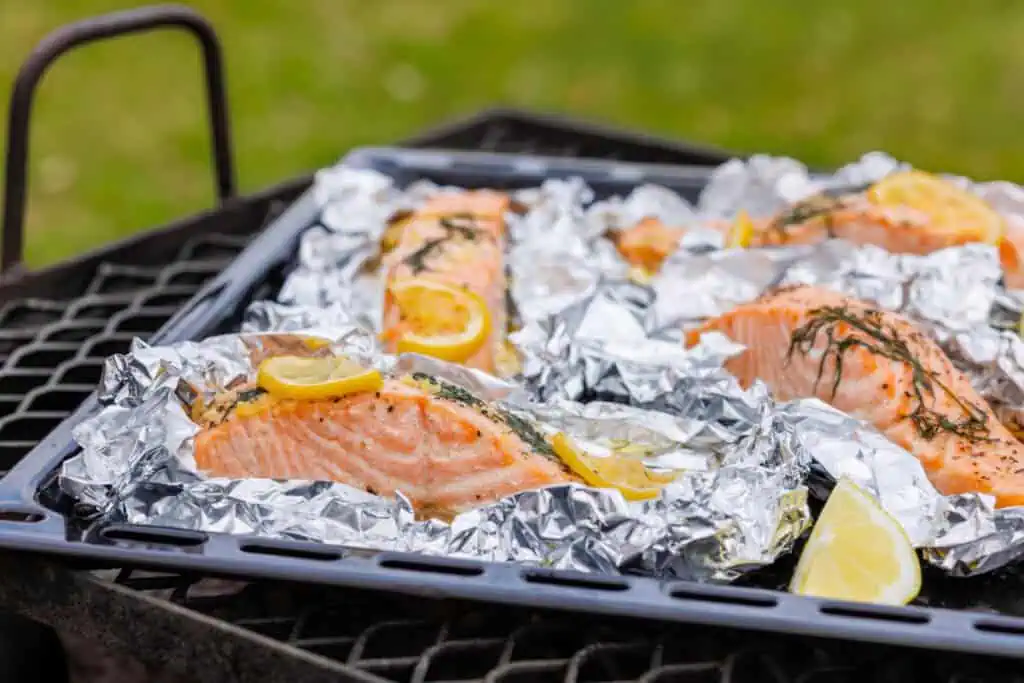 Four servings of lemon-dill salmon in foil packets, with lemon slices and dill on top, arranged on a black grill grate with a green grass background. One lemon wedge is in the foreground.