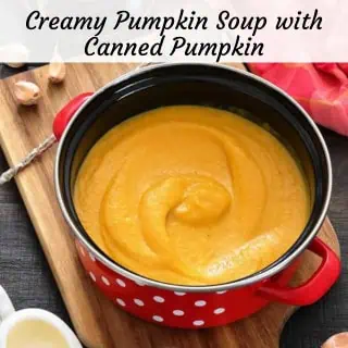 A red pot with white polka dots filled with creamy pumpkin soup sits on a wooden board, with garlic cloves and a white dish nearby.