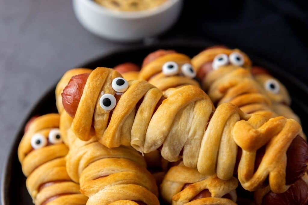 A plate of hot dogs wrapped in dough to look like mummies, with candy eyes on top. A bowl of dipping sauce is in the background.