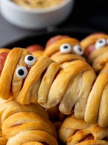 A plate of hot dogs wrapped in dough to look like mummies, with candy eyes on top. A bowl of dipping sauce is in the background.