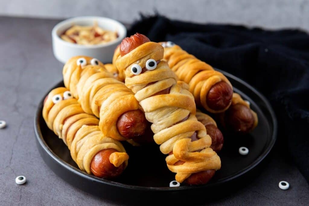 Hot dogs wrapped in golden-brown dough with candy eyes arranged on a black plate, resembling mummies, with a bowl of dipping sauce in the background.