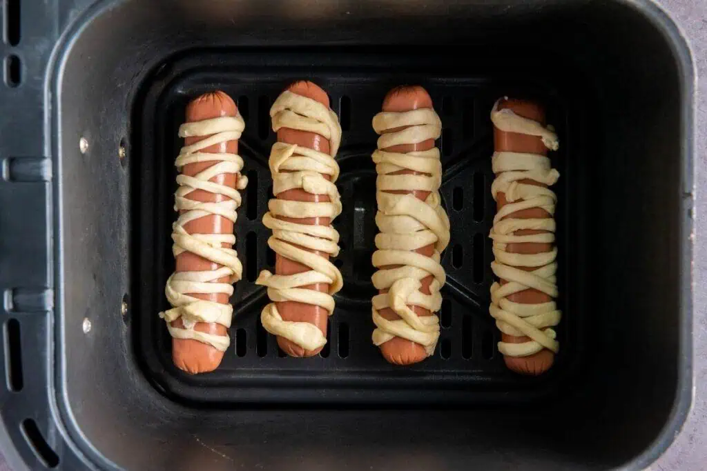Four hot dogs wrapped in strips of dough are arranged in a single layer inside an air fryer basket, ready to be cooked.