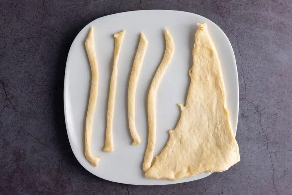 A white plate with several strips and one large piece of uncooked dough arranged on it, placed on a dark countertop.