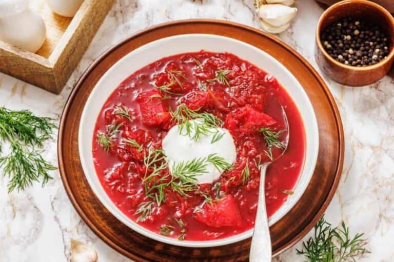 A bowl of red borscht soup with a dollop of sour cream and fresh dill on top, placed on a wooden plate with a spoon.