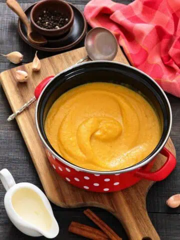 A red pot with creamy orange soup sits on a wooden board, surrounded by a ladle, garlic cloves, peppercorns, a pitcher of cream, and cinnamon sticks.