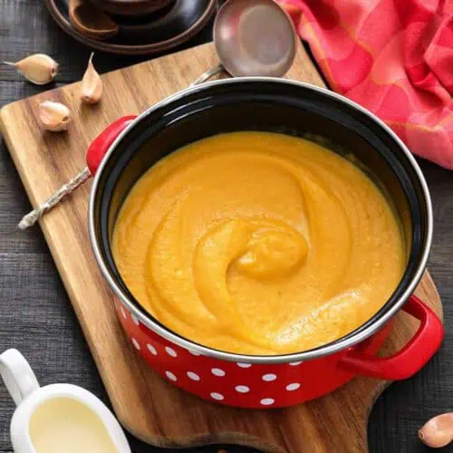 A red pot with creamy orange soup sits on a wooden board, surrounded by a ladle, garlic cloves, peppercorns, a pitcher of cream, and cinnamon sticks.