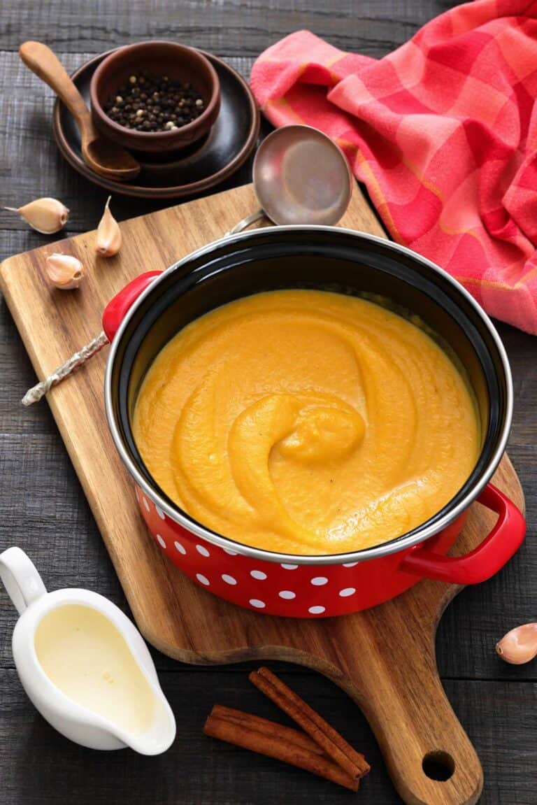 A red pot with creamy orange soup sits on a wooden board, surrounded by a ladle, garlic cloves, peppercorns, a pitcher of cream, and cinnamon sticks.