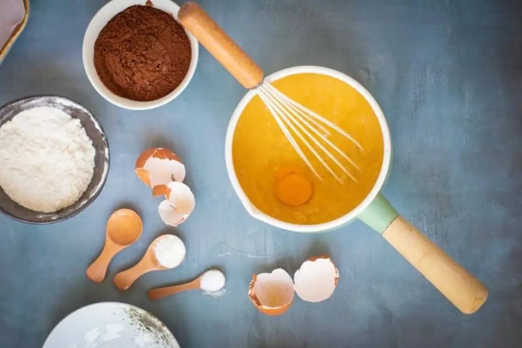 A mixing bowl with whisk, eggs, and batter, surrounded by cocoa powder, flour, egg shells, and measuring spoons on a blue surface.
