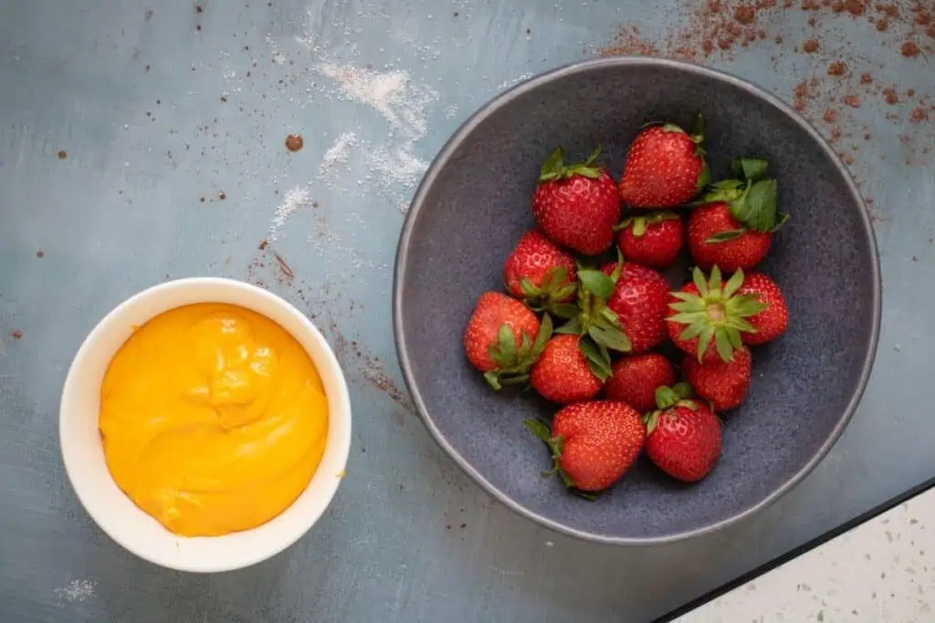 A gray bowl filled with fresh strawberries sits next to a white bowl containing yellow custard on a blue surface dusted with powder.