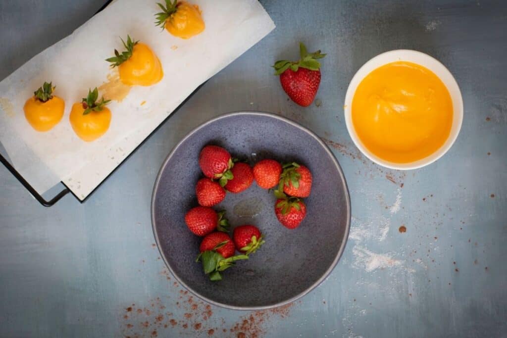 Fresh strawberries in a gray bowl, a white bowl of yellow melted coating, and parchment with strawberries partially coated in yellow on a blue surface.