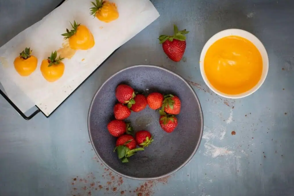 Fresh strawberries in a gray bowl, a white bowl of yellow melted coating, and parchment with strawberries partially coated in yellow on a blue surface.