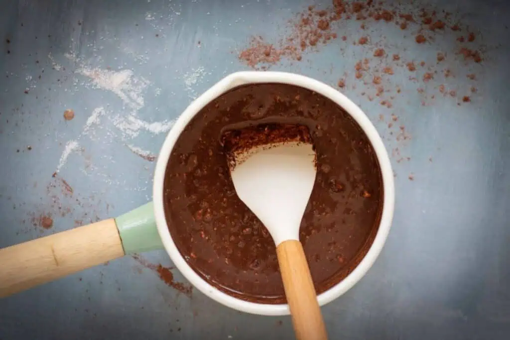 A white saucepan with a wooden-handled spatula contains chocolate batter, with cocoa powder scattered on a blue surface around it.