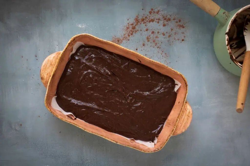 A rectangular baking dish filled with chocolate brownie batter sits on a gray surface next to a green saucepan with chocolate residue and a wooden spoon.