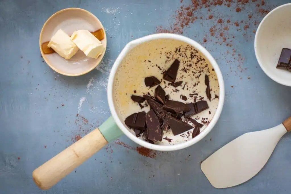 A saucepan with cream and chunks of dark chocolate, next to a plate with butter and a white spatula on a blue surface.