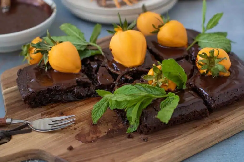 Sliced chocolate cake topped with glossy chocolate glaze, yellow fruit, and fresh mint leaves, served on a wooden board with a fork nearby.