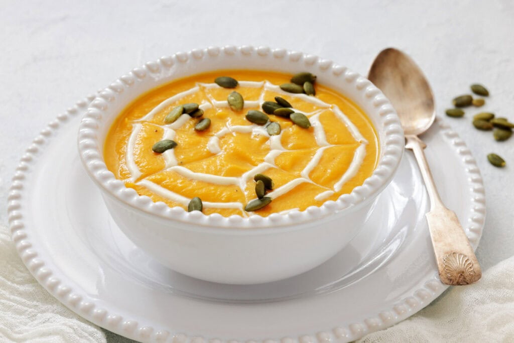 A white bowl of orange soup garnished with a swirl of cream and pumpkin seeds, served on a white plate with a vintage spoon.