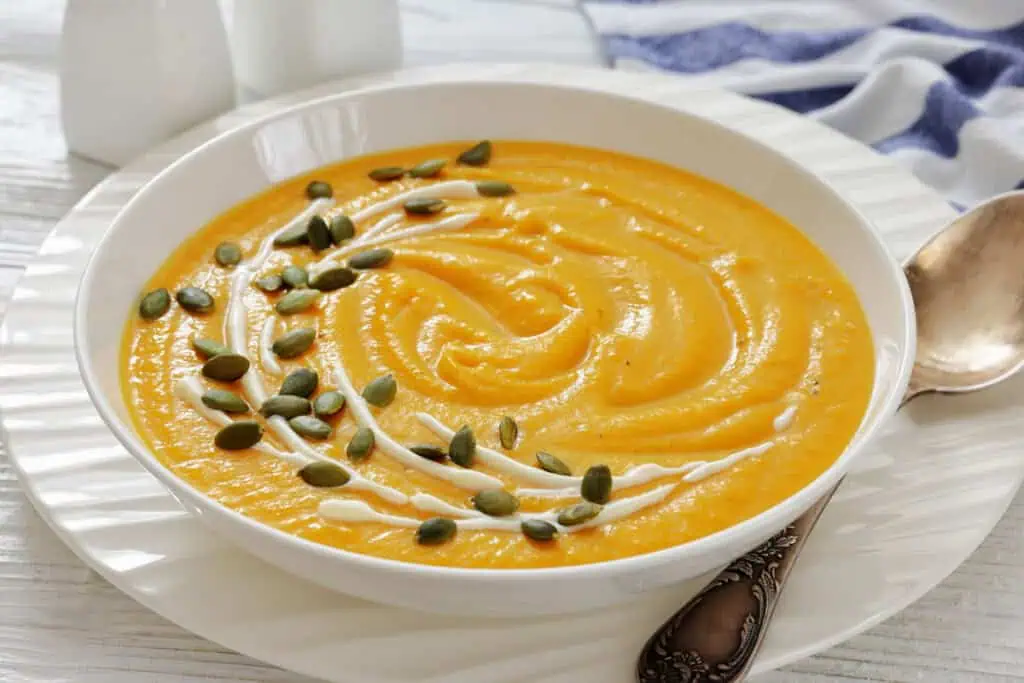 A bowl of creamy orange soup garnished with a swirl of cream and sprinkled with pumpkin seeds, placed on a white plate beside a spoon.