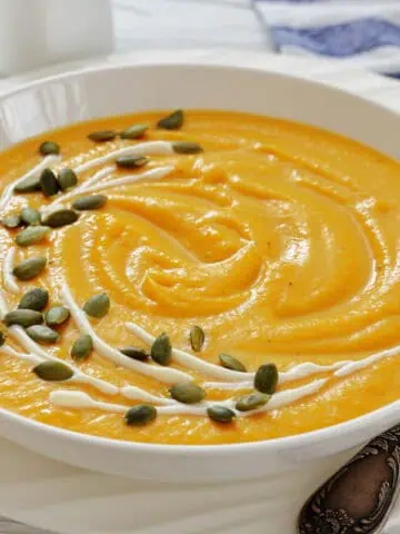 A bowl of creamy pumpkin soup with canned pumpkin garnished with a swirl of cream and sprinkled with pumpkin seeds, placed on a white plate beside a spoon.