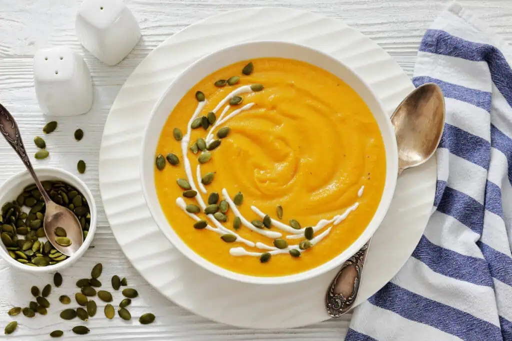 A bowl of creamy orange soup garnished with a swirl of cream and pumpkin seeds, served with a spoon on a white plate beside a striped napkin.