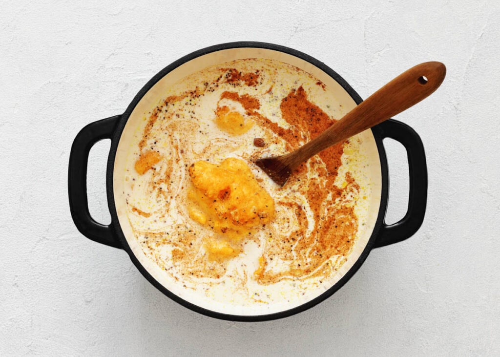 A black pot filled with cream, melted butter, and spices being mixed with a wooden spoon, on a white surface.