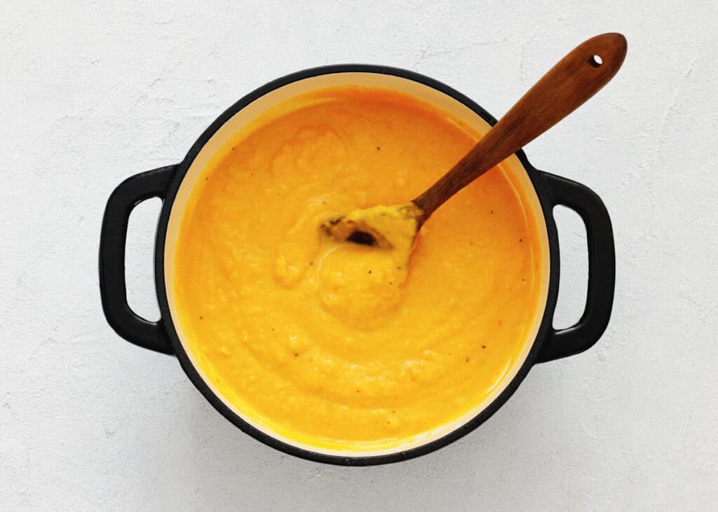 A pot of thick, creamy orange soup with a wooden spoon resting inside, viewed from above on a light surface.