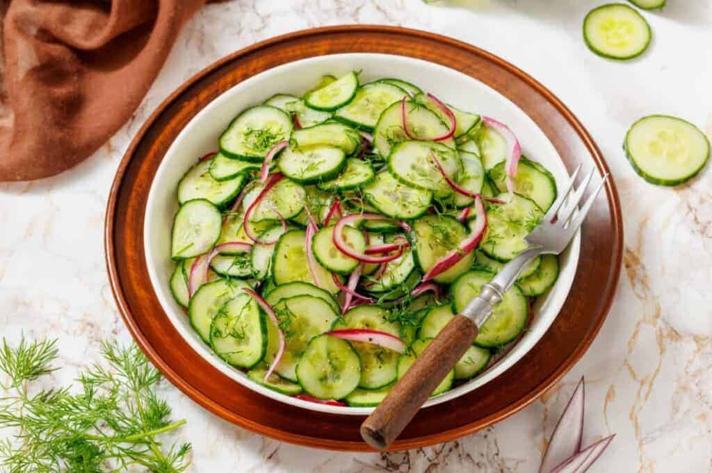 A bowl of cucumber salad with red onions and fresh dill, placed on a wooden plate with a fork, surrounded by ingredients on a marble surface.