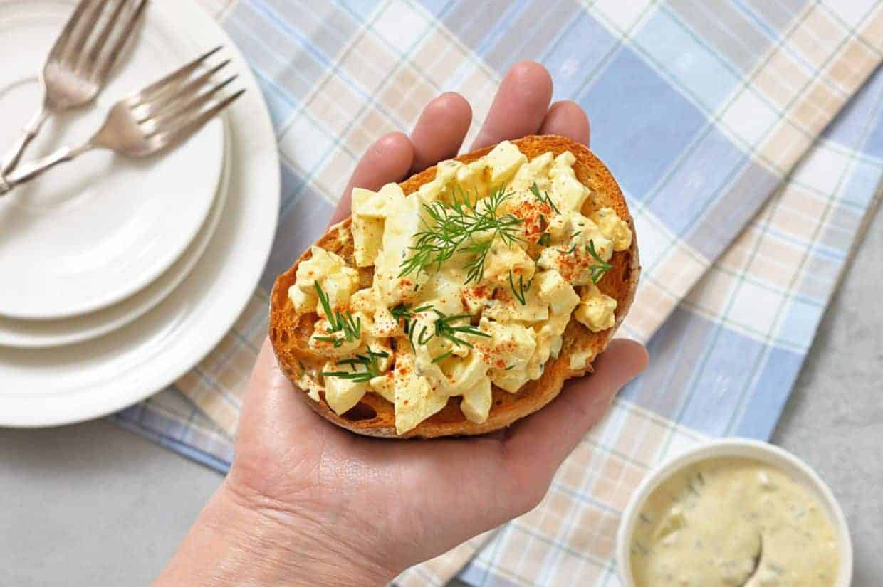 A hand holds a slice of Deviled Egg Toast, garnished with dill and paprika. Three stacked plates with forks and a bowl of sauce are in the background on a checked cloth.