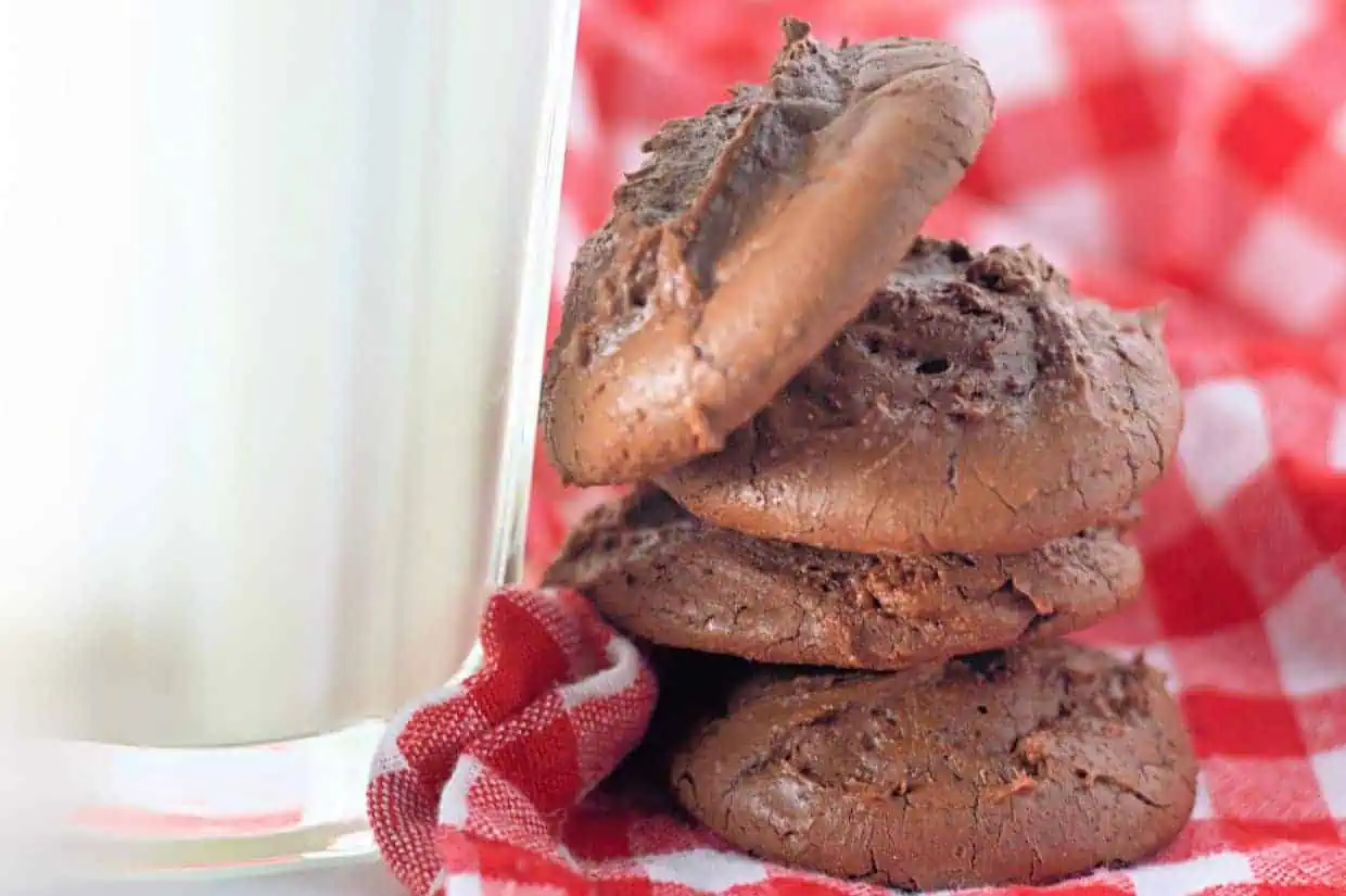 Three Fudgy Chocolate Brownie Cookies stacked beside a glass of milk on a red and white checkered cloth.