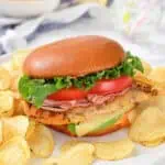A sandwich with lettuce, tomato, cheese, and sliced meat in a bun, served on a plate with potato chips.