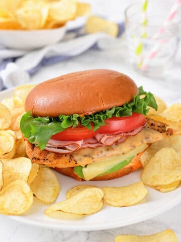 A sandwich with lettuce, tomato, cheese, and sliced meat in a bun, served on a plate with potato chips.