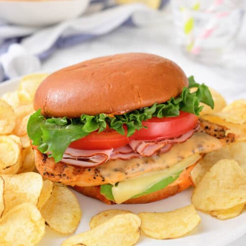 A sandwich with lettuce, tomato, cheese, and sliced meat in a bun, served on a plate with potato chips.