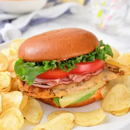 A sandwich with lettuce, tomato, cheese, and sliced meat in a bun, served on a plate with potato chips.
