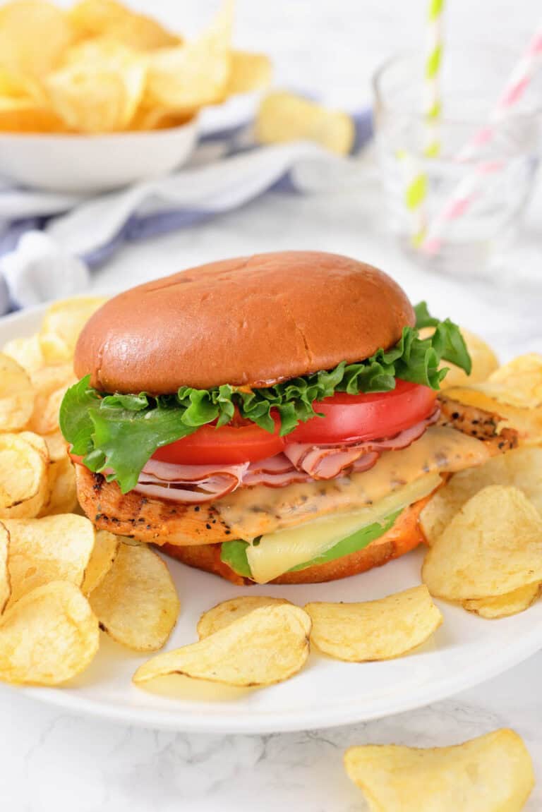 A sandwich with lettuce, tomato, cheese, and sliced meat in a bun, served on a plate with potato chips.