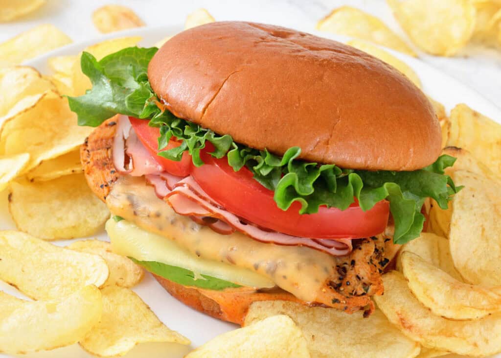 A grilled chicken sandwich with lettuce, tomato, ham, and cheese in a bun, served on a plate with potato chips around it.