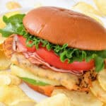 A grilled California chicken burger with lettuce, tomato, cheese, and ham on a bun, served with potato chips on a white plate.