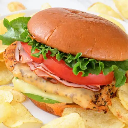 A grilled California chicken burger with lettuce, tomato, cheese, and ham on a bun, served with potato chips on a white plate.