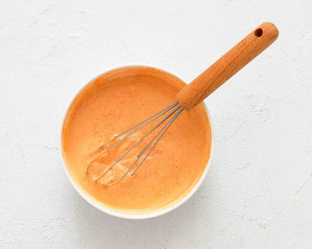 A whisk rests in a white bowl filled with an orange creamy sauce on a light textured surface.