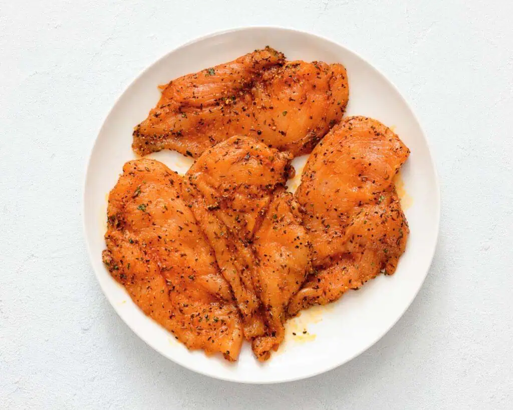 Four raw, seasoned chicken breasts are arranged on a white plate against a light background.