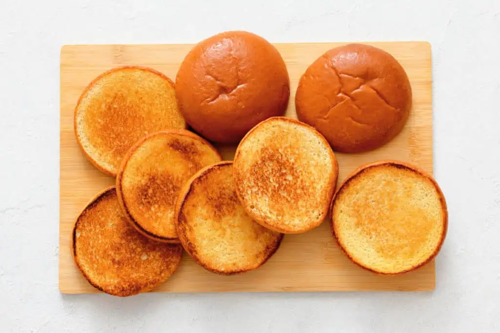 Four hamburger buns, with both tops and bottoms, are arranged on a wooden cutting board; the bottoms appear toasted while the tops are untoasted.