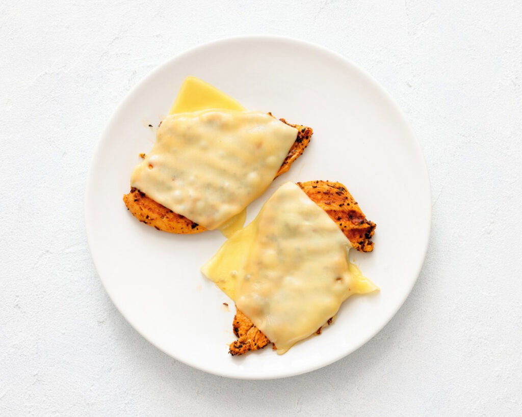 Two pieces of grilled chicken breast topped with melted cheese slices on a white plate.