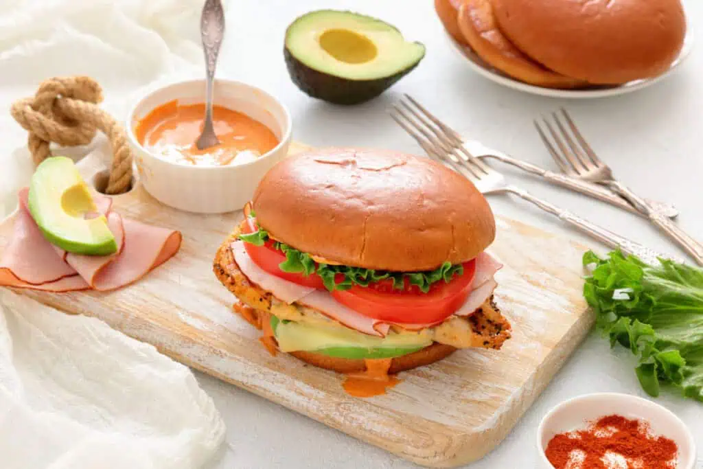 A grilled chicken sandwich with ham, lettuce, tomato, avocado, and sauce on a brioche bun sits on a cutting board, with extra sauce, avocado, and utensils nearby.