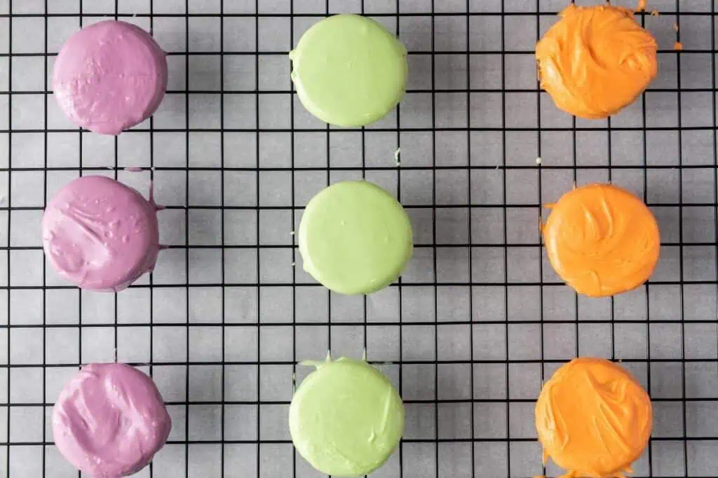 Nine round, coated cookies in purple, green, and orange sit on a black cooling rack placed over parchment paper.